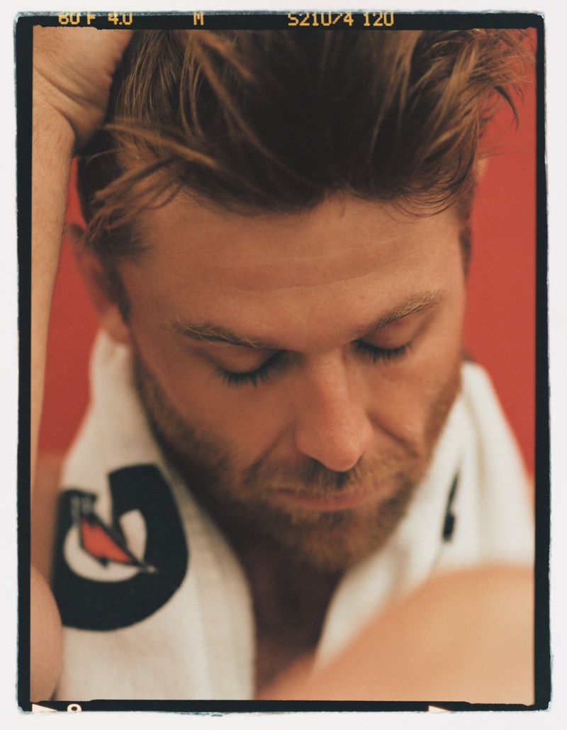 Close-up portrait of a fair-skinned adult man with short brown hair, a light beard, eyes closed, wearing a towel around his neck, against a red background.