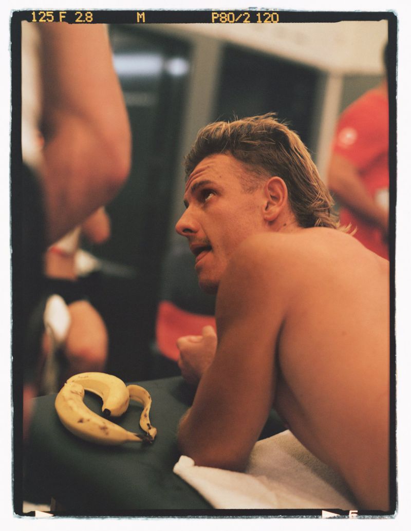 Blonde man with a mullet hairstyle lies topless on a table next to a peeled banana in a gym-like indoor setting, shallow depth of field.