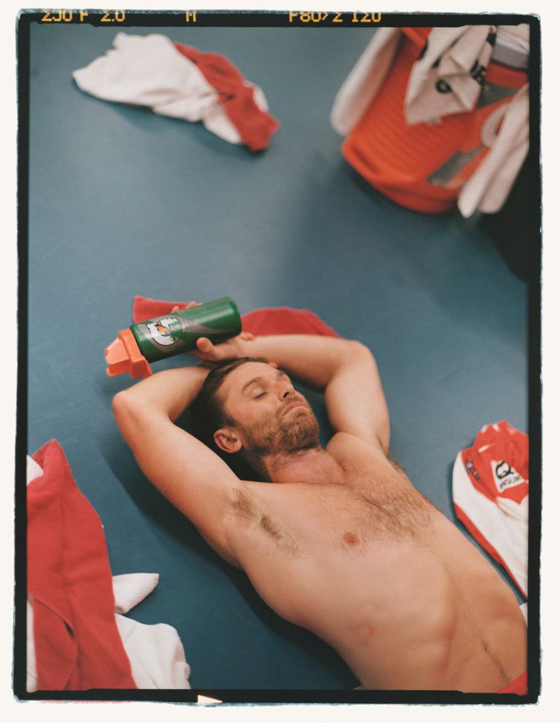 Shirtless man lying on floor with energy drink, arms behind head in locker room with clothing; athletic, fitness, and relaxation themes.