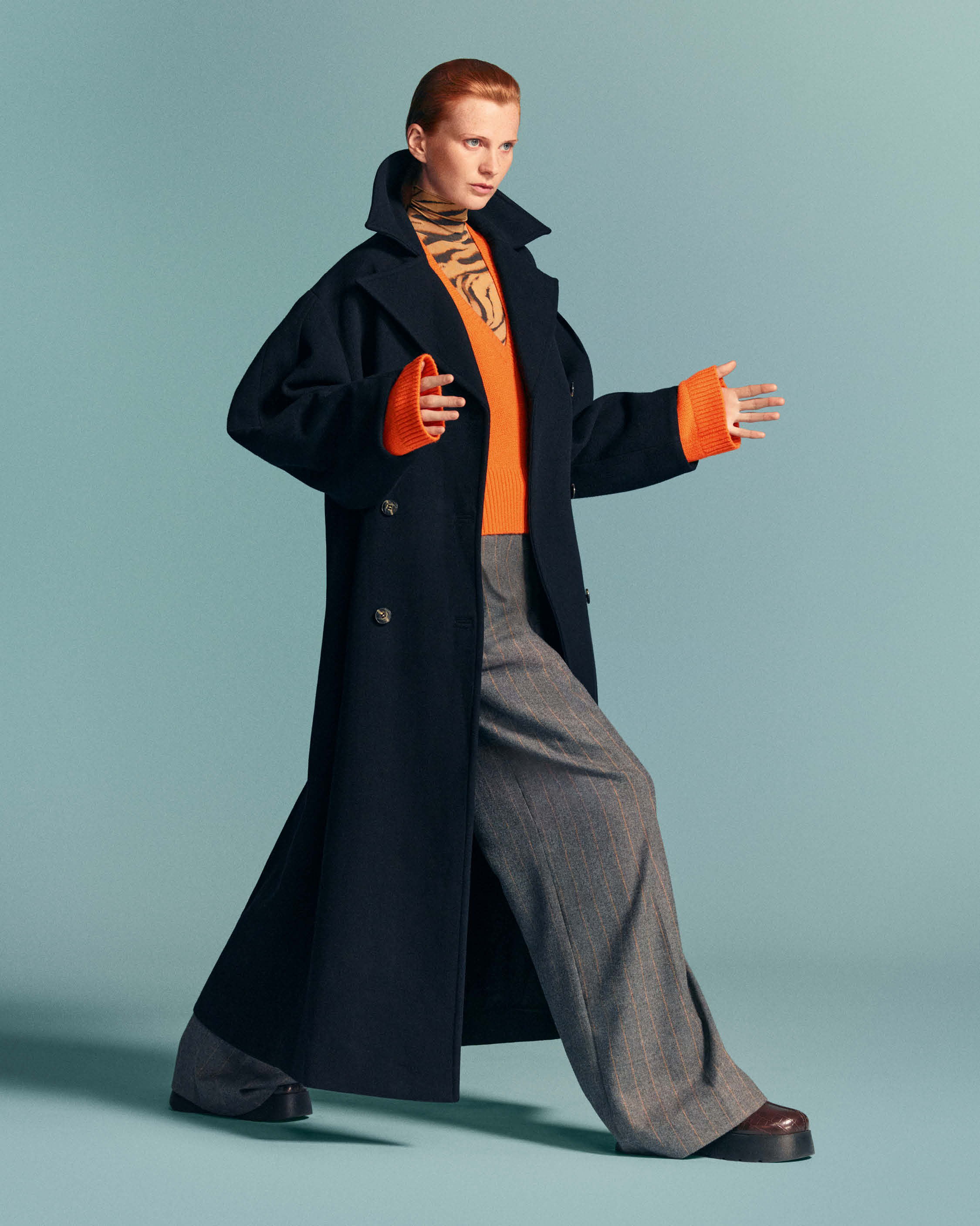 Model wearing a long dark blue coat over an orange sweater and grey trousers against a solid turquoise backdrop.