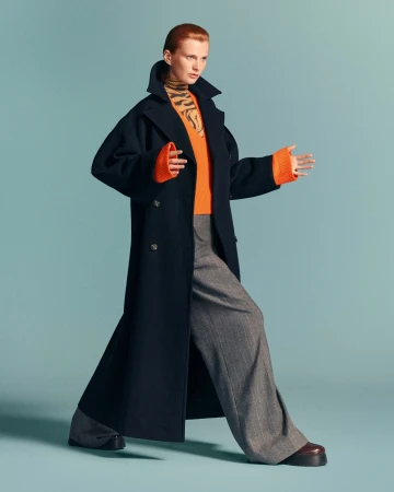 Model wearing a long dark blue coat over an orange sweater and grey trousers against a solid turquoise backdrop.