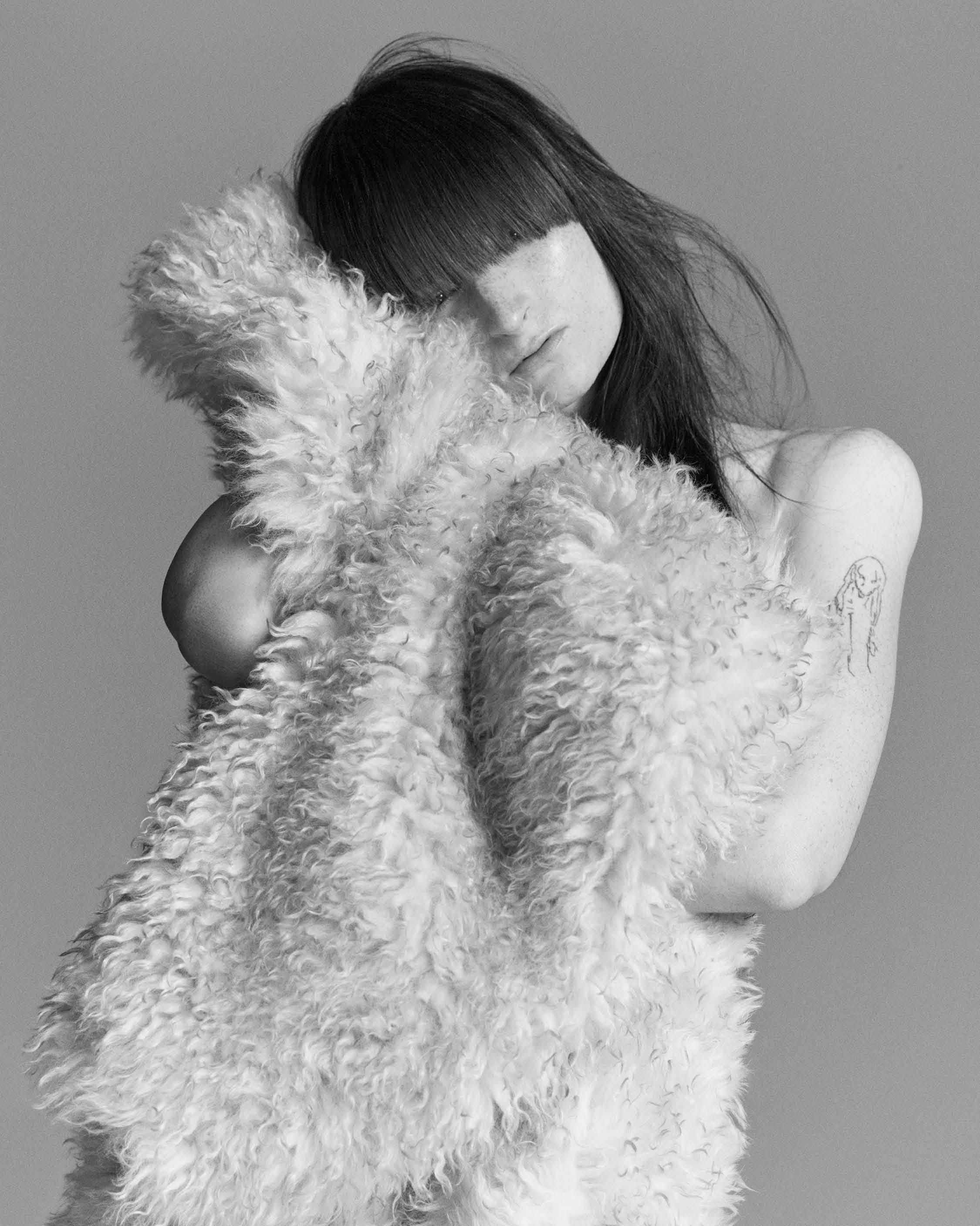 Model with long dark bangs embracing a fluffy textured garment against a plain grey backdrop