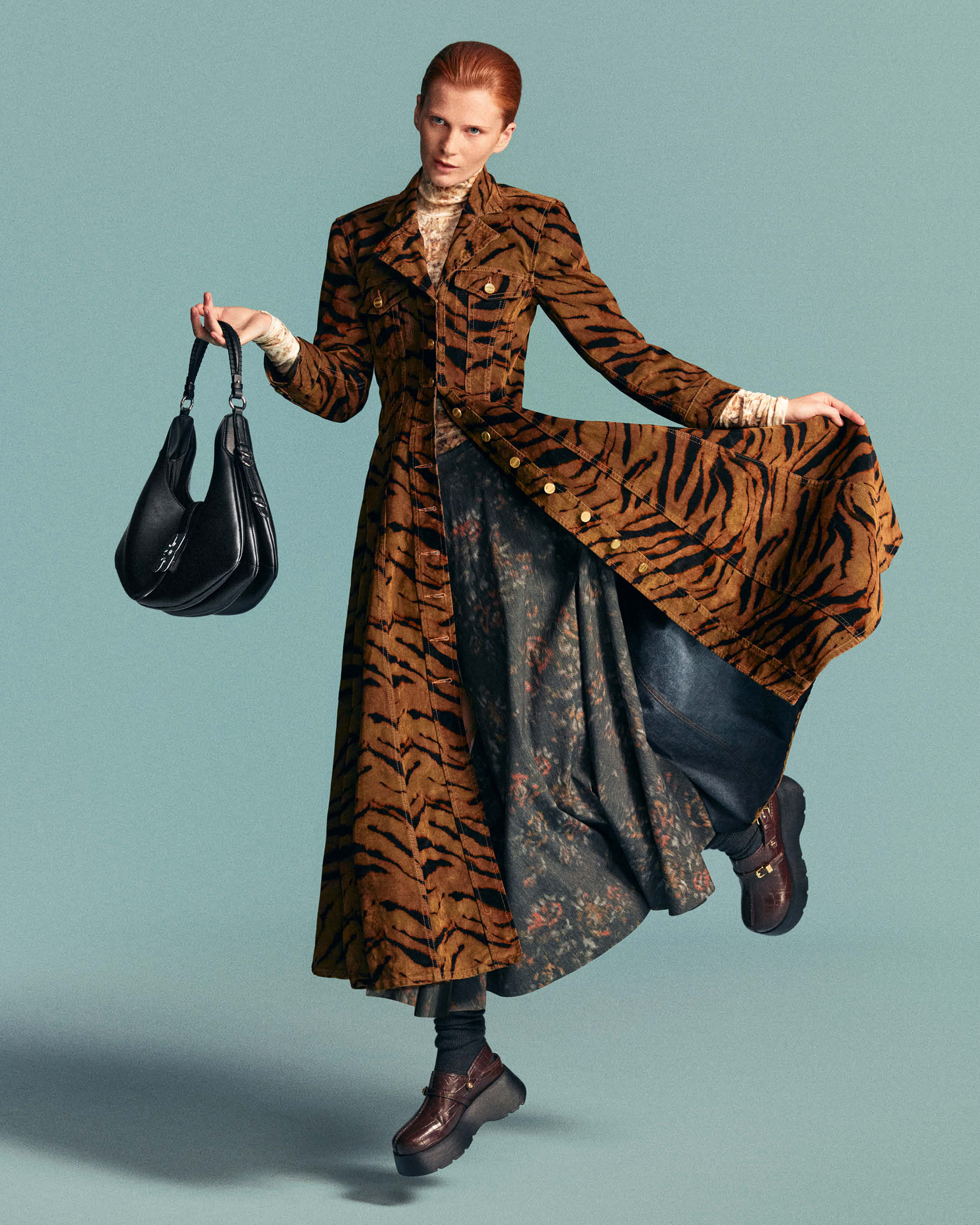 Model in a brown tiger-stripe coat holding a black handbag against a solid light blue background