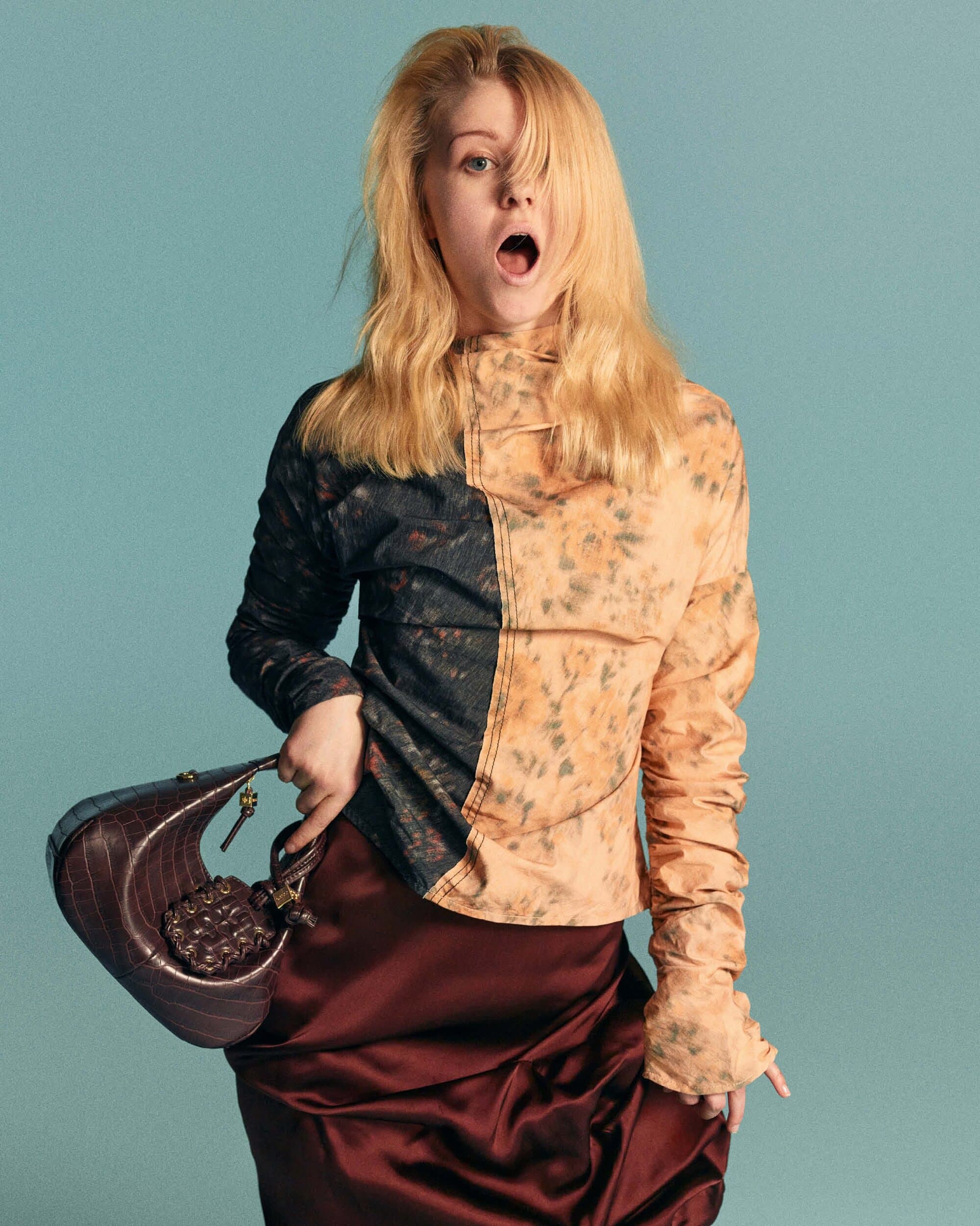 Model with long blonde hair wearing a patterned top and holding a dark handbag against a turquoise background