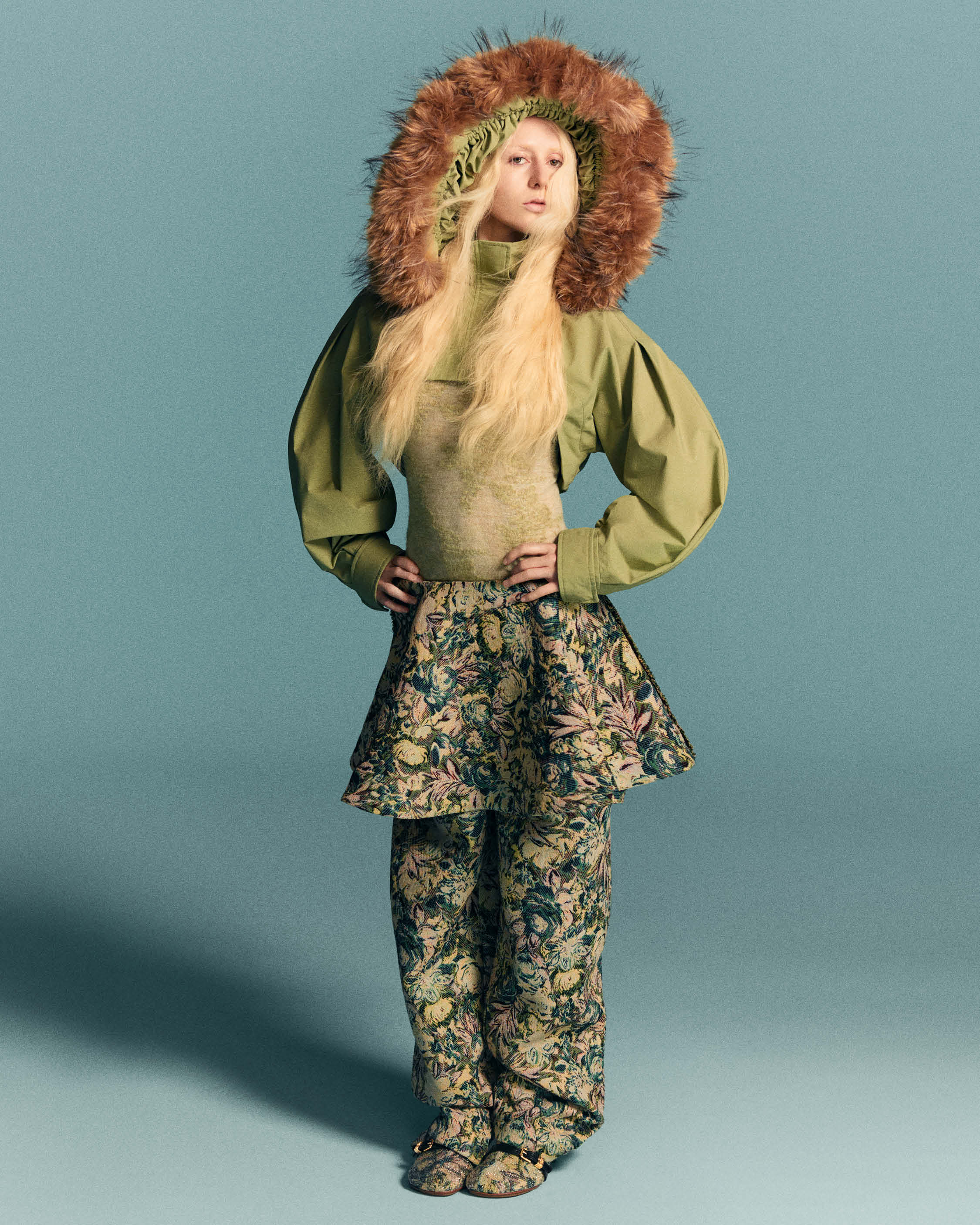 Model wearing a green top with a furry hood and patterned matching trousers against a solid background