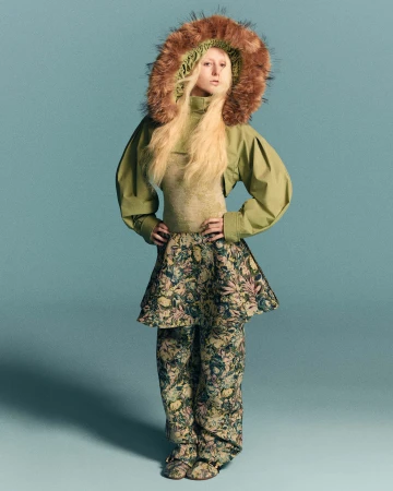 Model wearing a green top with a furry hood and patterned matching trousers against a solid background