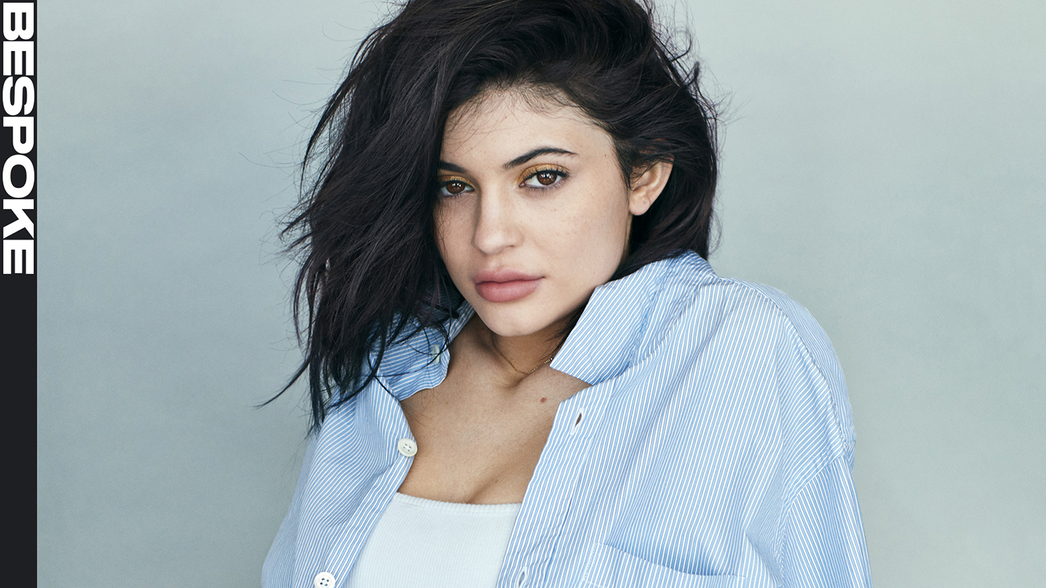 Kylie Jenner wearing a blue striped button-down shirt against a light grey studio backdrop with vertical Bespoke text on the left.