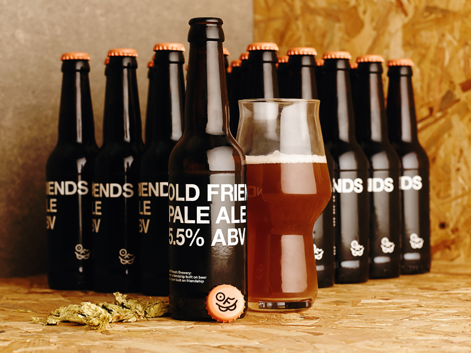 Beer bottles and a filled glass of pale ale with a bottle cap resting on a wooden chipboard surface