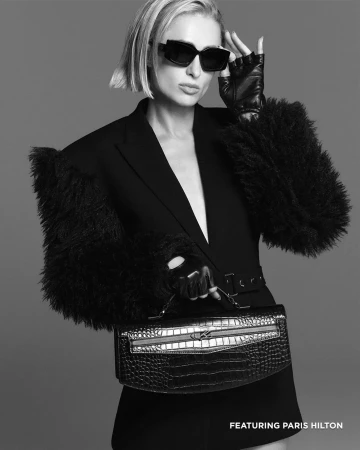Paris Hilton wearing black sunglasses and a dark blazer with furry sleeves holding a handbag against a grey background