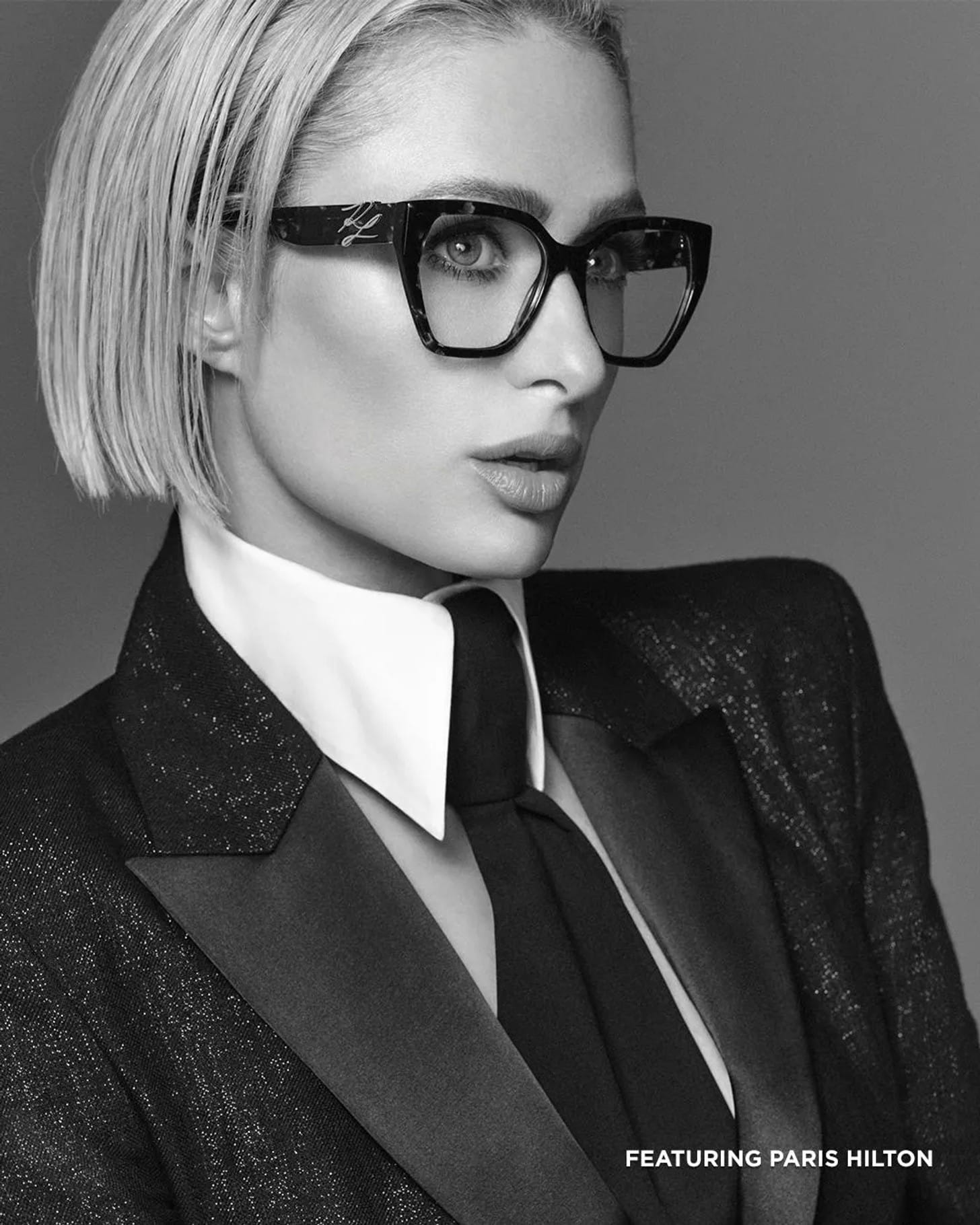 Black and white portrait of Paris Hilton wearing dark geometric eyeglasses, a crisp white collared shirt, black tie, and a tailored suit jacket.