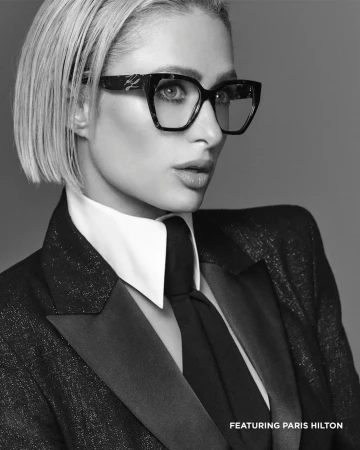 Black and white portrait of Paris Hilton wearing dark geometric eyeglasses, a crisp white collared shirt, black tie, and a tailored suit jacket.