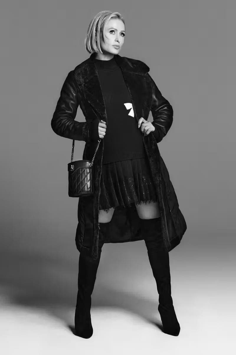 Paris Hilton in a black coat and boots holding a quilted handbag against a grey seamless studio background.