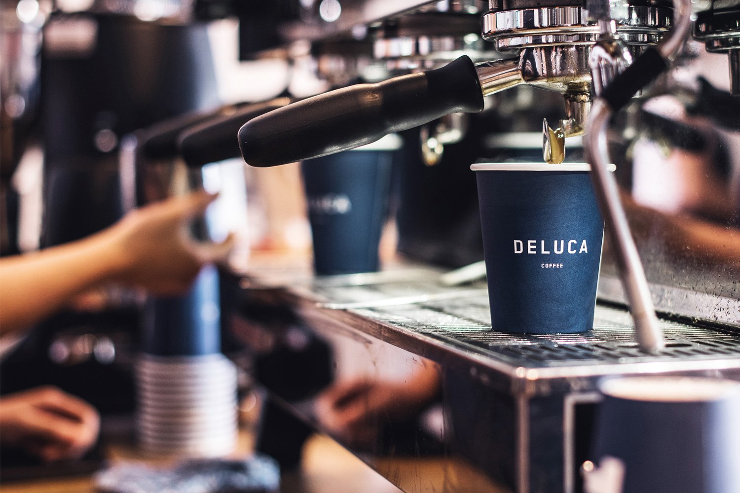Deluca Coffee paper cup filled under an espresso machine in a cafe setting