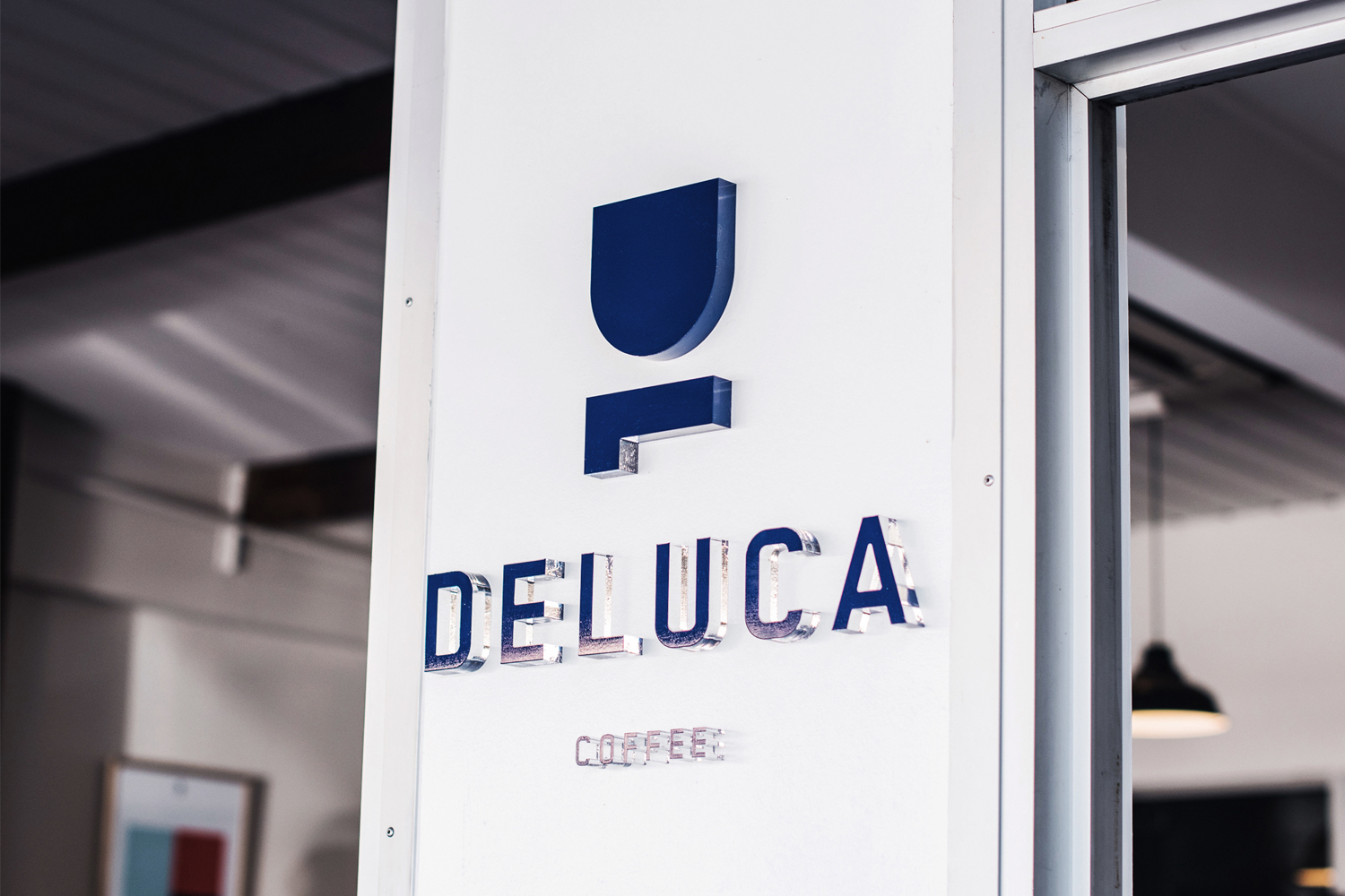 Dimensional blue acrylic signage spelling Deluca Coffee mounted on a white exterior wall next to a glass doorway.