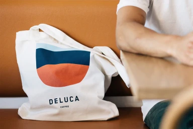 White canvas tote bag featuring a modern graphic and Deluca Coffee branding, resting on a brown leather bench next to a seated person.