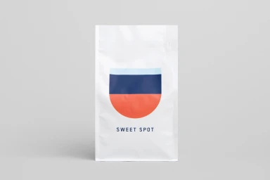 White Deluca Coffee pouch featuring a minimal geometric logo with blue and orange stripes on a grey background.