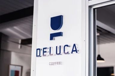 Dimensional blue acrylic signage spelling Deluca Coffee mounted on a white exterior wall next to a glass doorway.