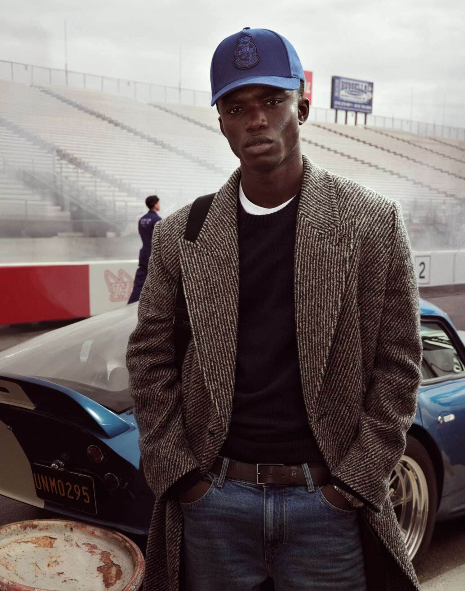 Black man wearing a blue cap, tweed coat, and jeans standing in front of a blue car and a racing stadium background