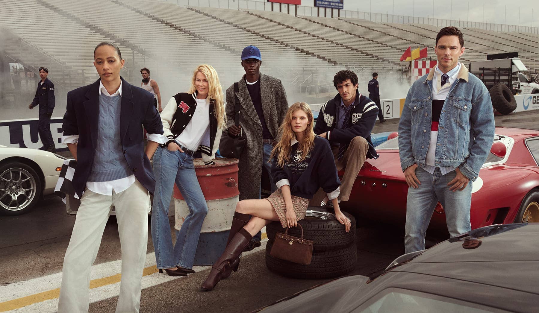 Group of six models wearing preppy apparel posing at a racetrack with classic race cars and bleachers background