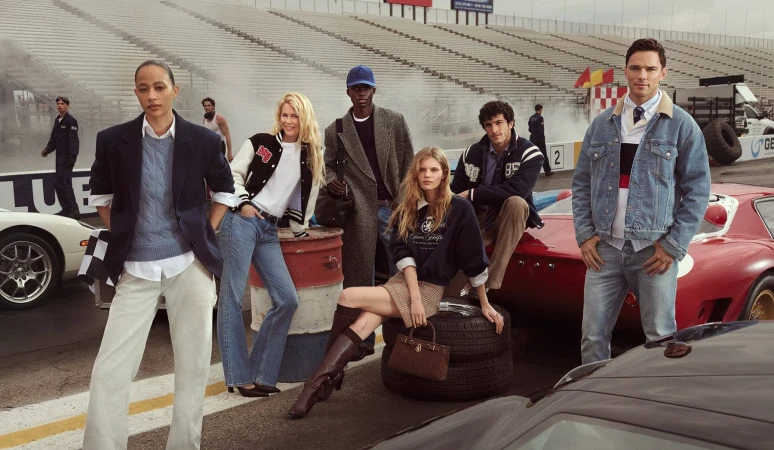 Group of six models wearing preppy apparel posing at a racetrack with classic race cars and bleachers background
