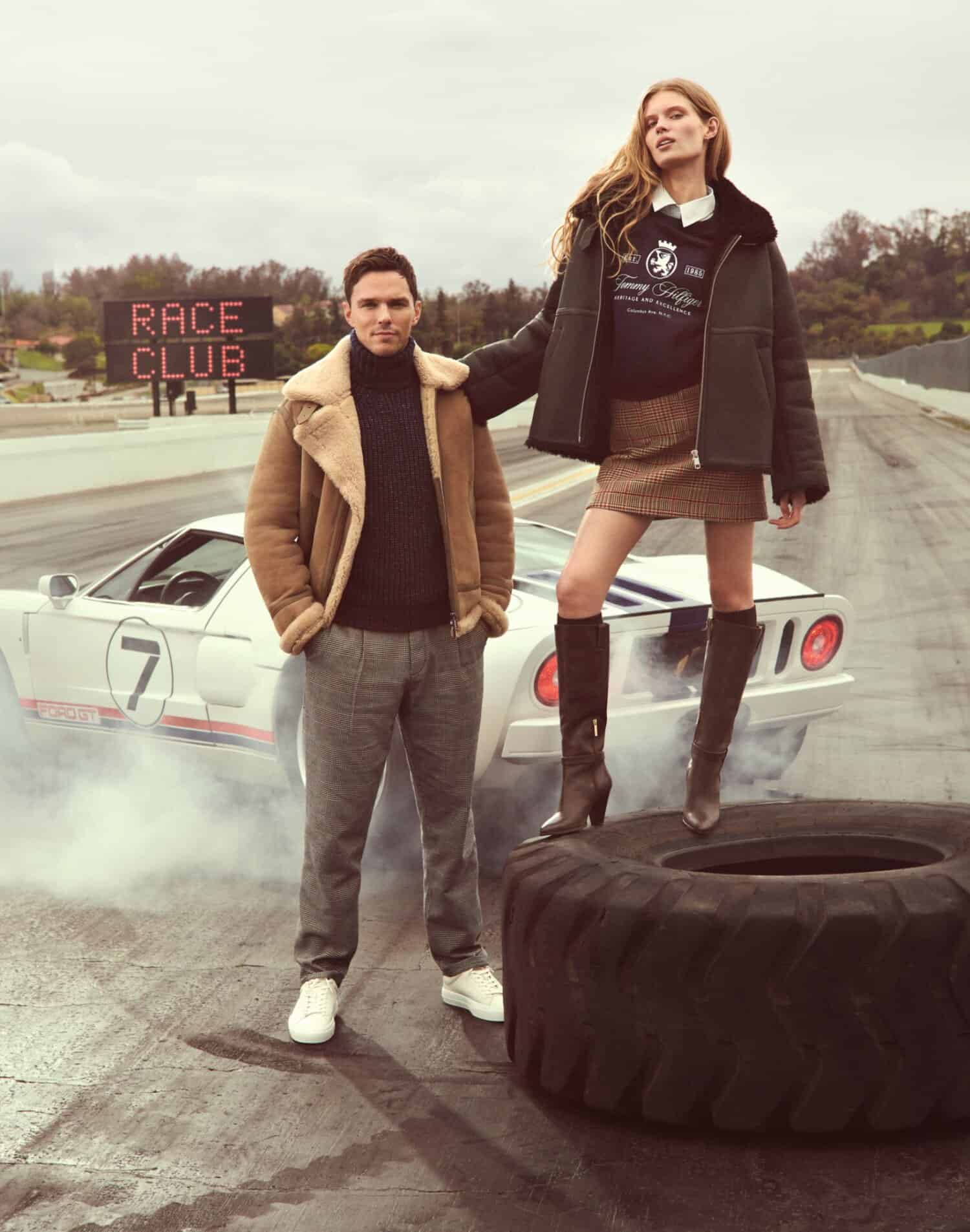 Man standing on asphalt next to a white sports car while a woman poses on a large tire with smoke rising around them.