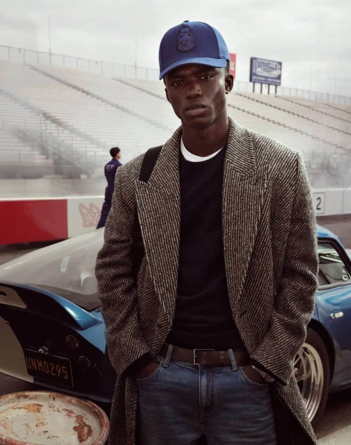 Black man wearing a blue cap, tweed coat, and jeans standing in front of a blue car and a racing stadium background