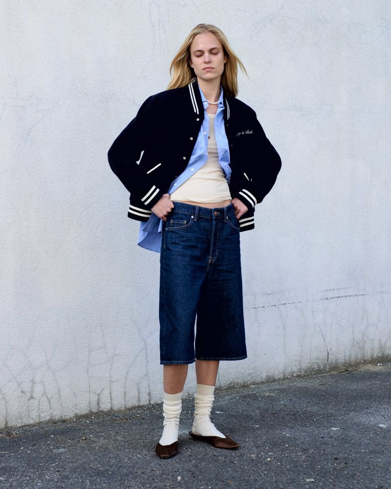 Fashion shot of a woman wearing an open blue shirt, dark denim shorts, jacket and flats, posing against a textured grey background.