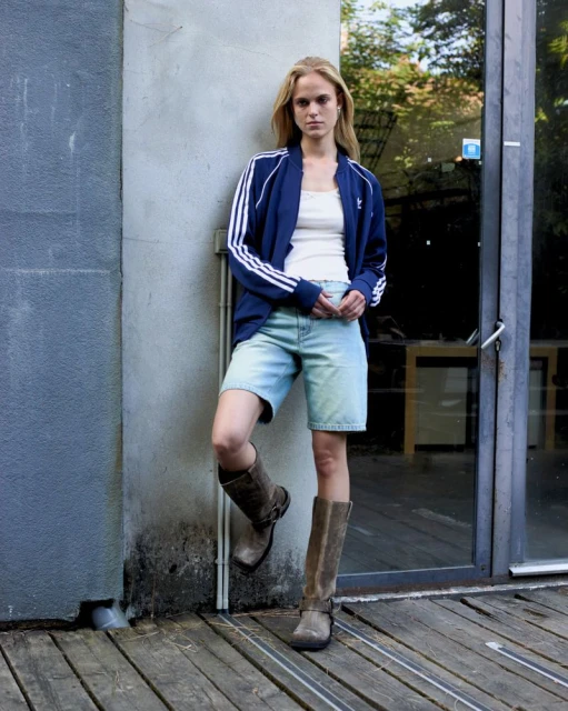 Woman in blue Adidas track jacket, light denim shorts, and knee-high boots stands against a gray wall with a glass door backdrop.