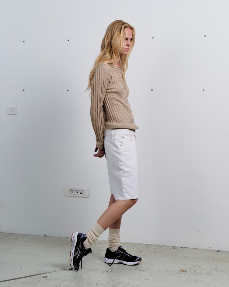 Blonde woman poses in a neutral outfit with a beige sweater, white skirt, socks, and running shoes against a blank white wall in studio.