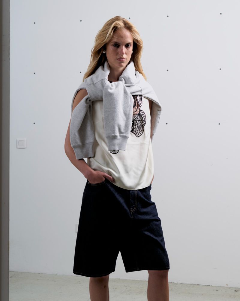 Fashionable young adult female model in white tank top and dark denim shorts posing in a neutral-toned studio environment with natural lighting.