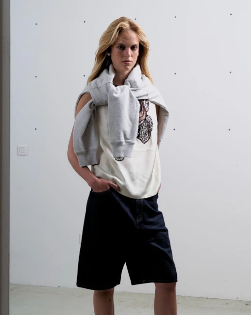 Fashionable young adult female model in white tank top and dark denim shorts posing in a neutral-toned studio environment with natural lighting.