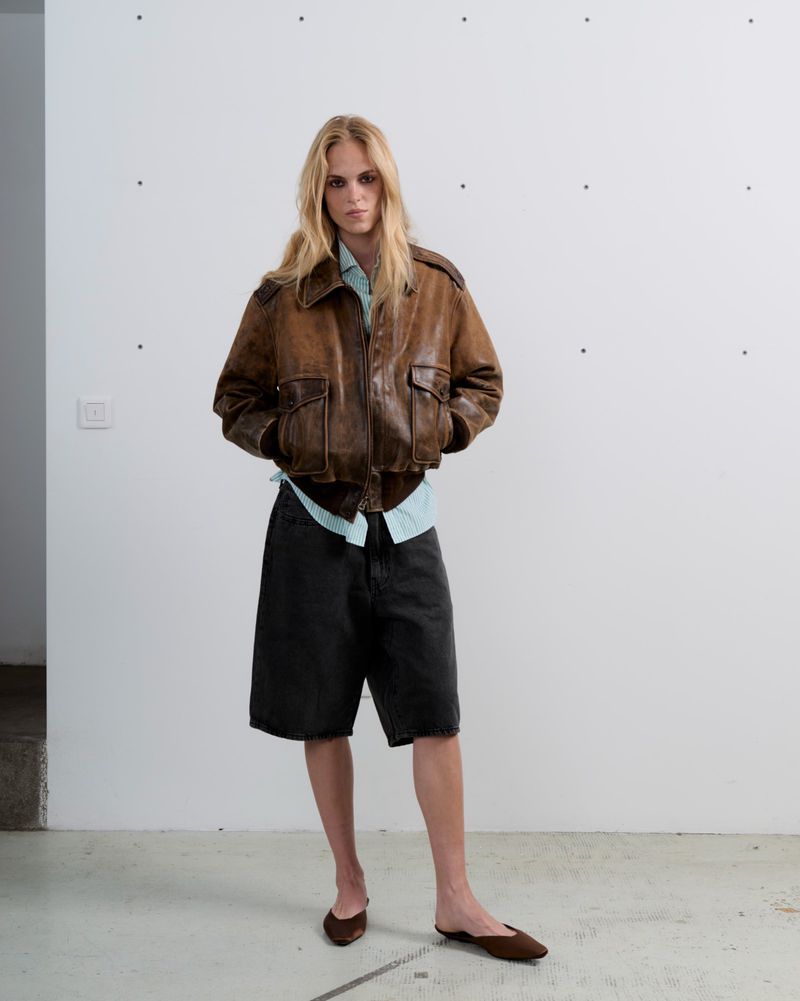 A slender adult poses in an indoor photo studio, wearing a brown leather bomber jacket, dark denim shorts, and brown flats against a white wall.