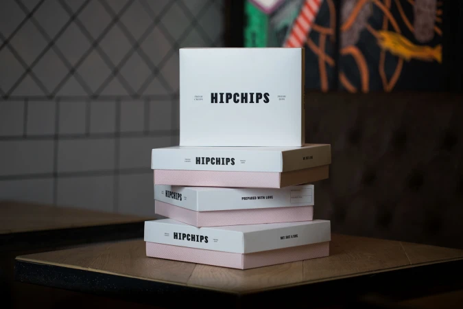 A stack of white and pink Hipchips branded boxes resting on a wooden table in a dimly lit interior.