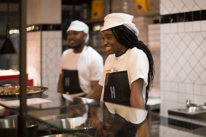 Two smiling kitchen staff wearing white caps, white t-shirts, and dark aprons behind a service counter at Hipchips.