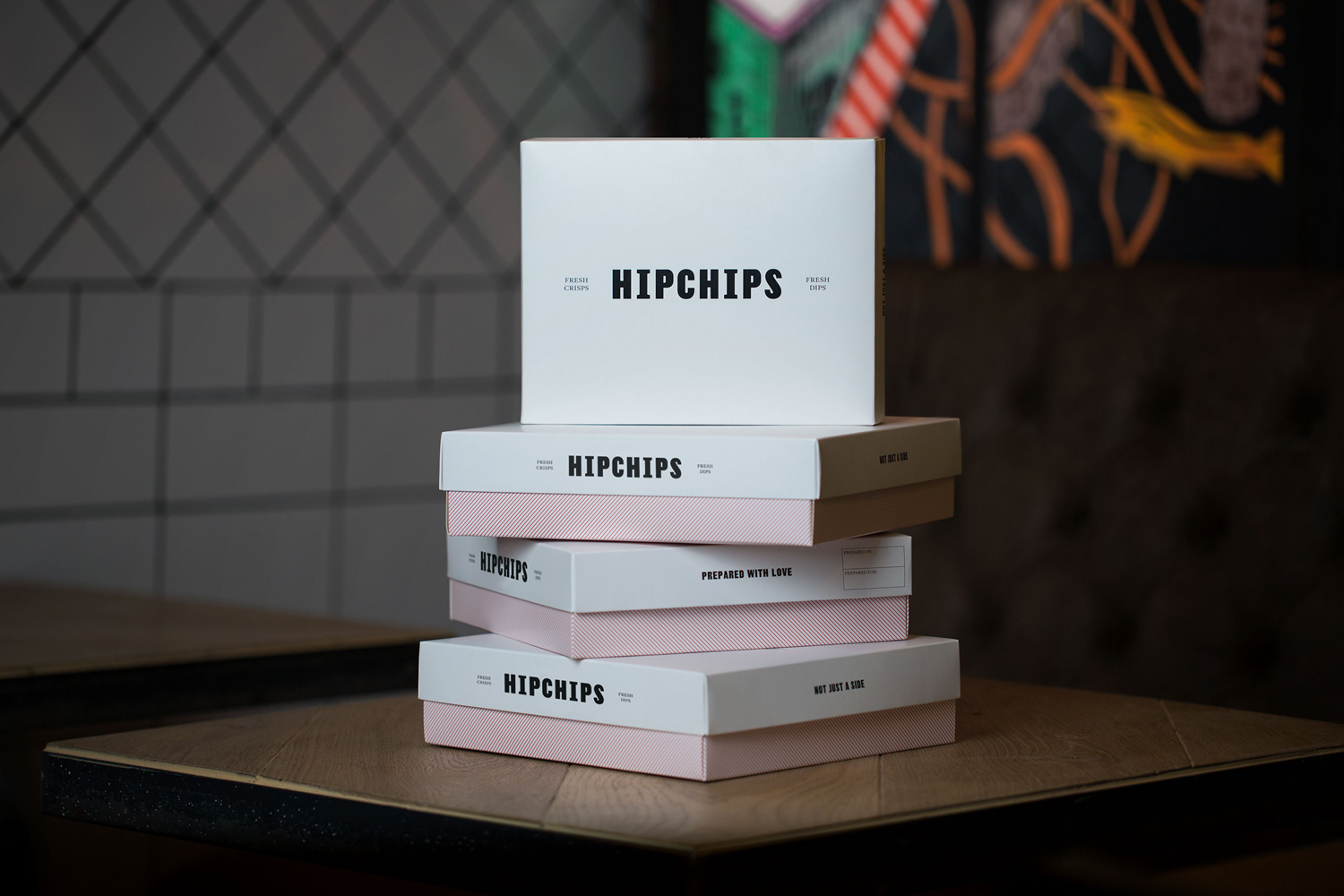 A stack of white and pink Hipchips branded boxes resting on a wooden table in a dimly lit interior.