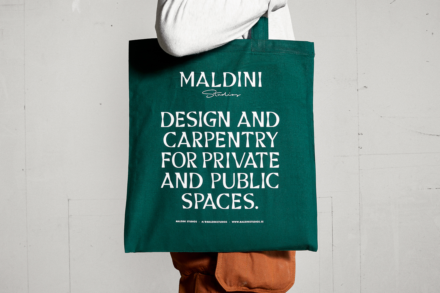 Person holding a dark green canvas tote bag with white typography against a neutral concrete background