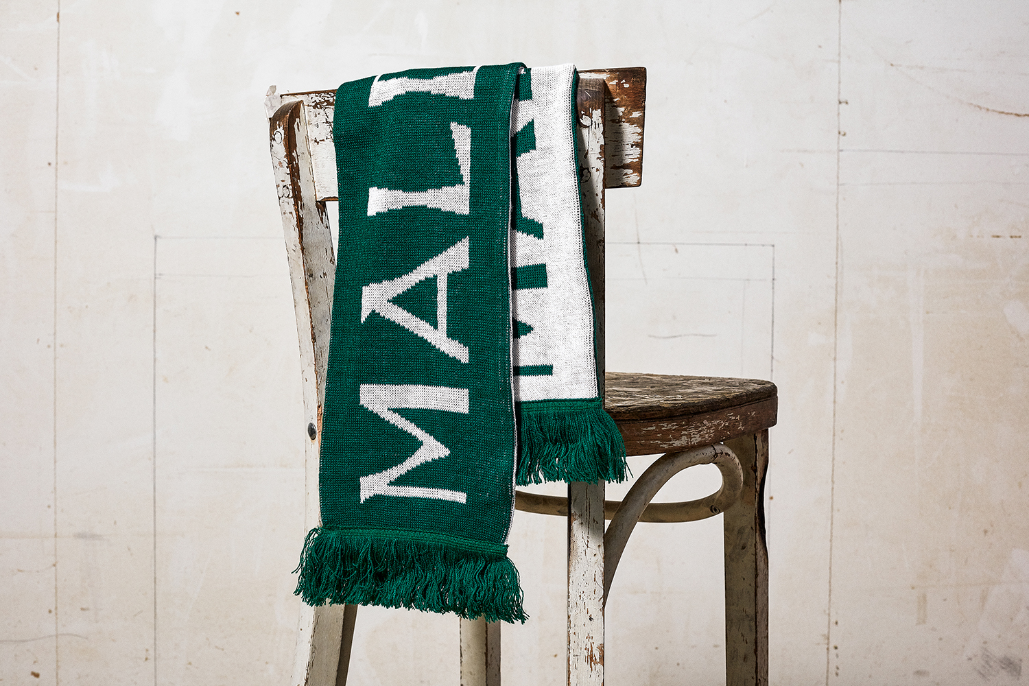 Green and white knitted scarf draped over a rustic distressed wooden chair against a neutral paneled wall background.