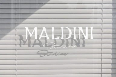 Maldini Studios brand identity typography overlaid on horizontal window blinds with soft diagonal shadows.