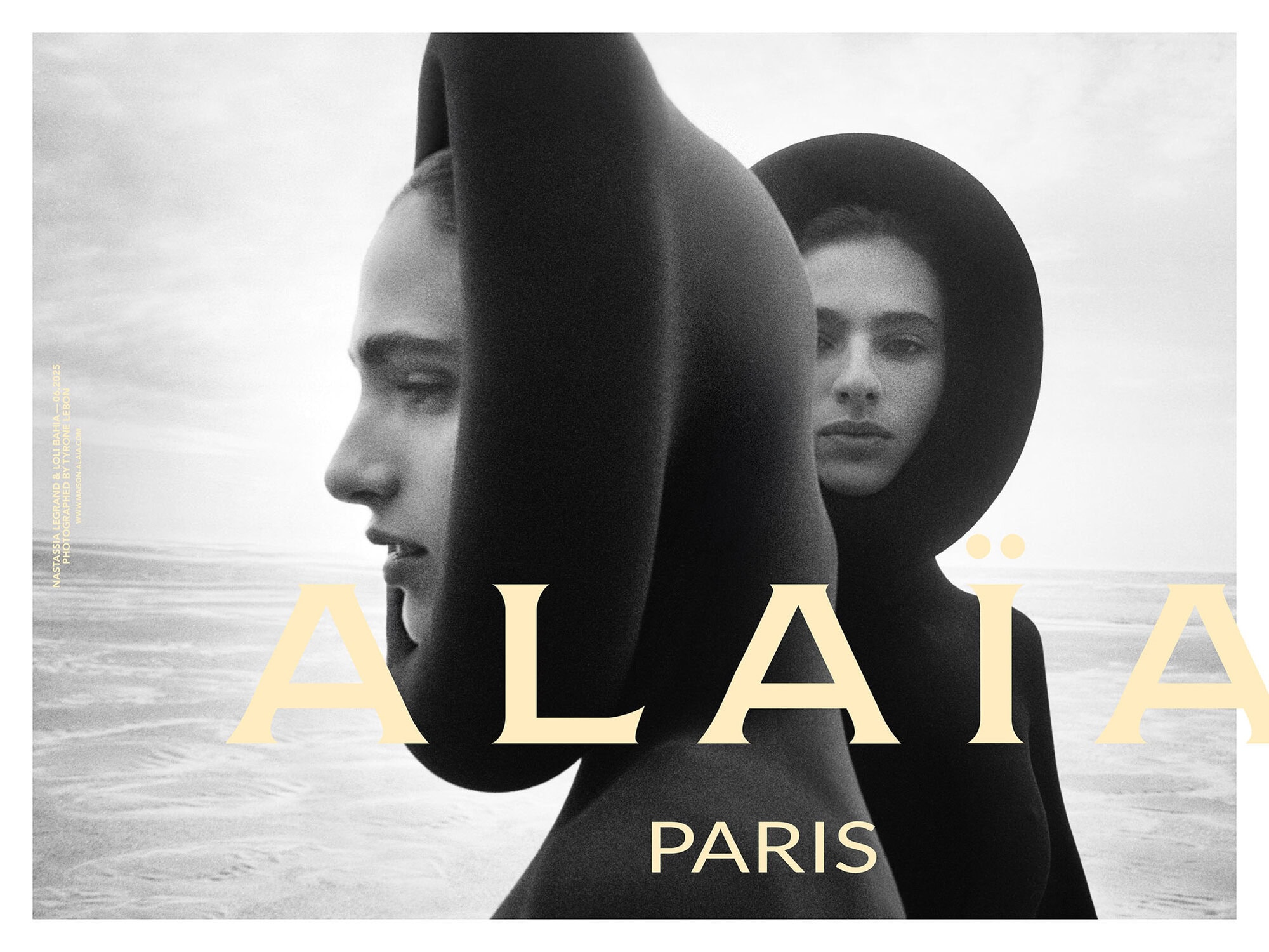 Two female models wearing black sculptural hooded garments outdoors against an overcast sky and coastal background.