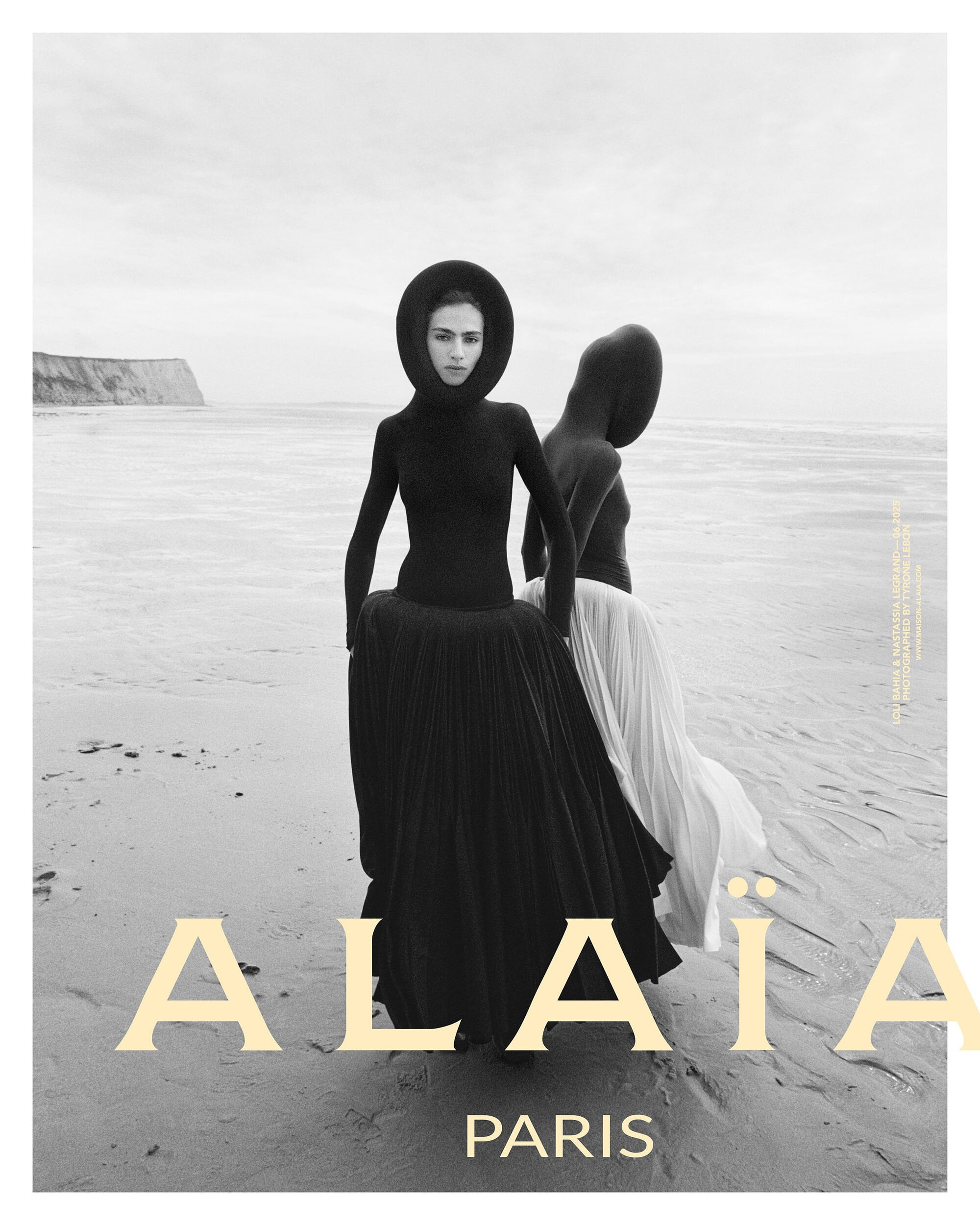 Two models in dark sculptural hooded garments standing on a sandy beach under an overcast sky for Alaia Fall 2025 campaign.