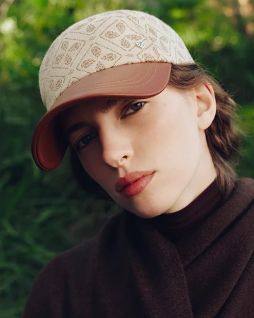 Close-up portrait of a woman wearing a patterned cap with a brown brim and a dark brown sweater against a green background