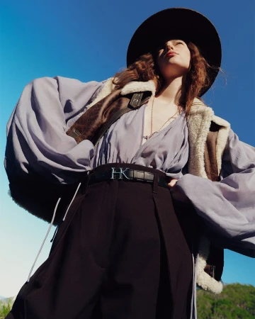 Low-angle shot of a model wearing a wide-brimmed hat, shearling jacket, and black trousers against a clear blue sky