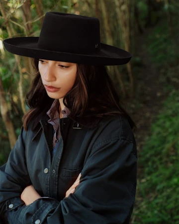 Woman wearing a wide-brim black hat and dark jacket outdoors against a natural green forest background