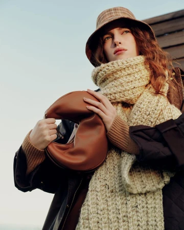 Model wearing a plaid bucket hat and chunky knit scarf holding a brown leather handbag outdoors.