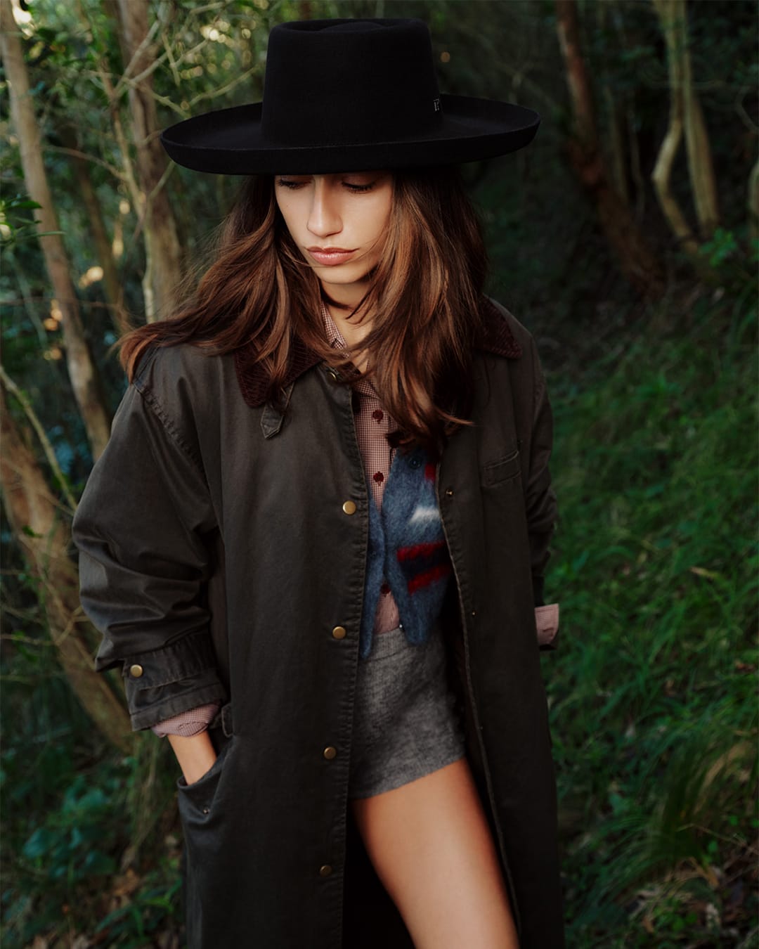 Model wearing a black wide-brim hat and dark green coat standing outdoors in a dense forest setting