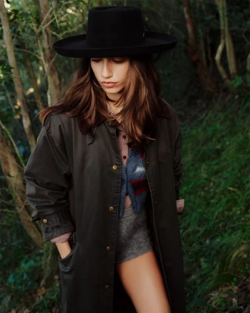 Model wearing a black wide-brim hat and dark green coat standing outdoors in a dense forest setting