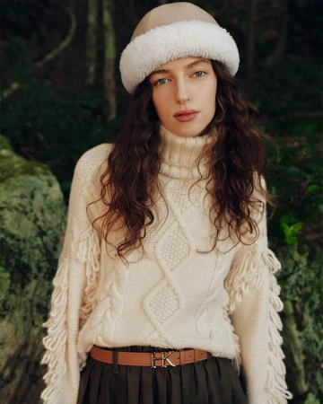 Model wearing a cream turtleneck sweater with fringe detailing and a white shearling hat outdoors
