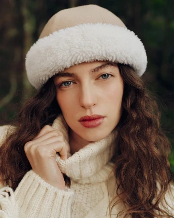 Model wearing a beige hat with white faux-fur trim and a cream knit sweater against an outdoor green backdrop.
