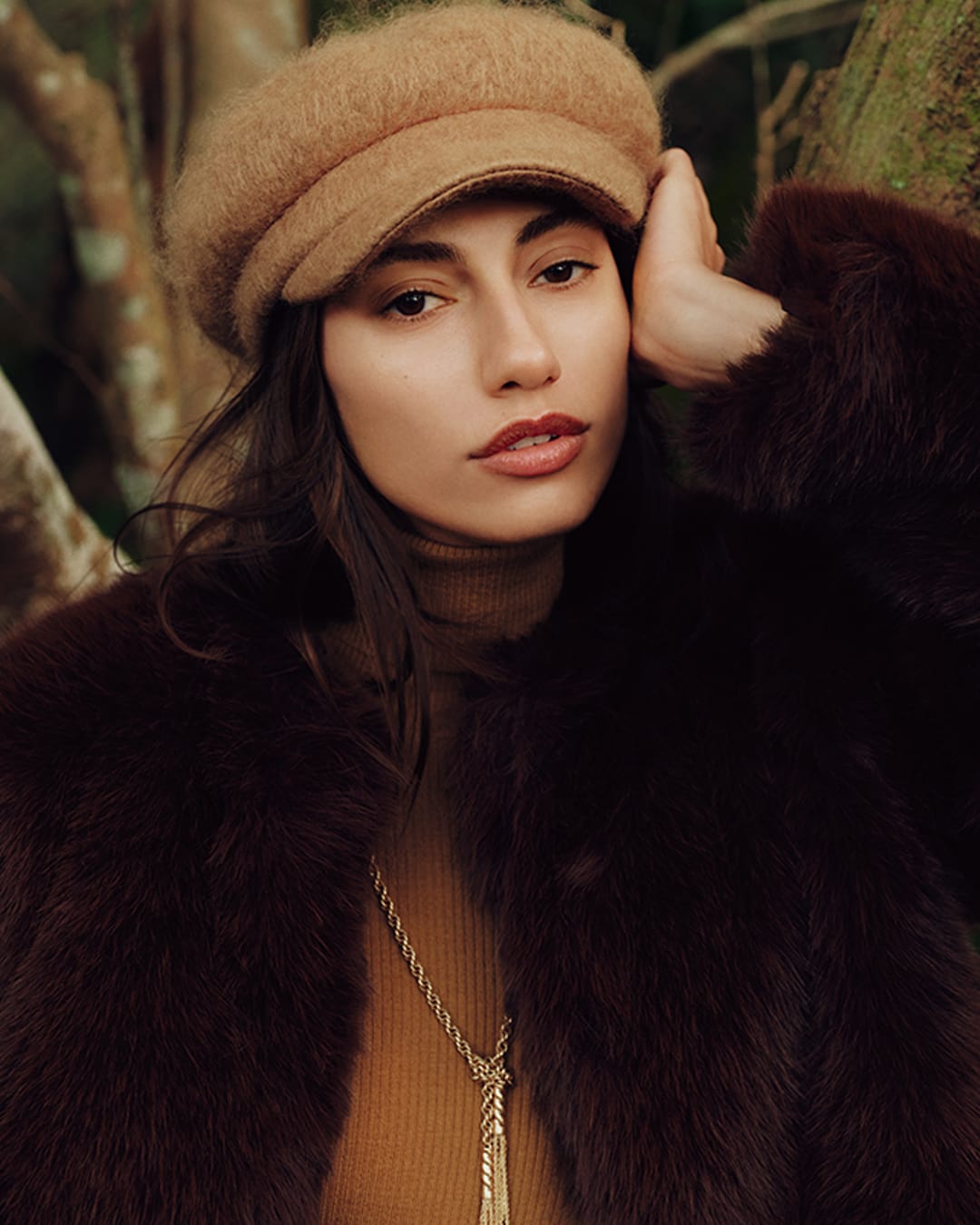 Model wearing a brown cap and dark furry coat with a gold necklace against a natural background
