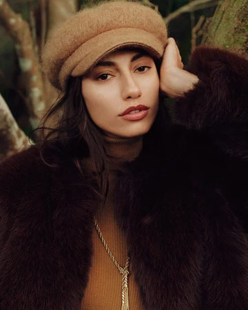 Model wearing a brown cap and dark furry coat with a gold necklace against a natural background